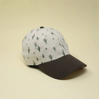 Storied Hats Adult Hat, Cactus Sunset, is made from organic cotton with a white crown featuring small cactus prints and a solid black brim. Its side profile evokes a Grand Canyon sunset when placed on a light beige surface.