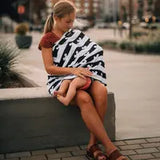 A woman sits on a concrete bench, using a Milk Snob Cover Disneys Mickey Mouse to breastfeed her child. Blurred buildings and plants line the city street in the background. Shes wearing a red top and sandals, seamlessly blending into the urban scene.