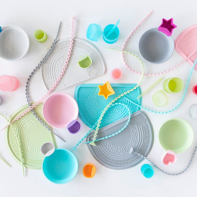 Flatlay image of Bumkins Silicone Grip Bowls, Sensory Placemats, Accessory Tethers, Cups, and Little Dippers styled on a white table.