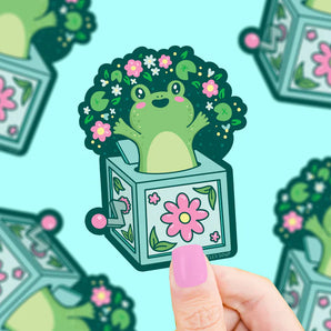 Turtle Soups Vinyl Sticker, Frog Jack In The Box features a green frog with a flowery afro emerging from a floral Jack-in-the-box. The background shows the repeating pattern, while the foreground displays this charming weatherproof sticker held in a hand.