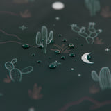 Close-up of water droplets on Bumkins dark green, waterproof fabric with a desert theme, featuring cacti, stars, and a crescent moon. This whimsical night-time aesthetic fabric is perfect for the Reusable Snack Bag, Large 2 Pack: Desert Night and is machine washable.