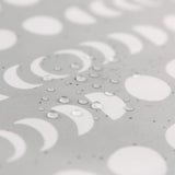 The Bumkins Wet Bag: Lunar Phase features a close-up of gray waterproof fabric adorned with crescent and full moon patterns. Water droplets highlight the textured material and heat-sealed seams.