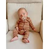 A baby in a SIIX Collection Organic Romper, Saddle Stripe—featuring fold-over mitten and foot cuffs—sits on a white chair with bare feet against the cushioning. A pacifier rests in its mouth as it looks slightly to the side.