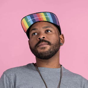 A bearded person in a Storied Hats Pride hat and gray shirt looks up against a pink backdrop. The hat reads Love is Love is Love.