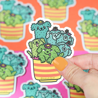 A hand with painted nails holds Turtle Soups premium vinyl sticker, Cactus Pups, showcasing cartoon cacti shaped like dogs in a colorful pot. It decorates a backdrop of similar waterproof stickers on vibrant orange and pink surfaces.