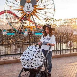 A woman in a Mickey Mouse shirt smiles, pushing a stroller with a Milk Snob Cover Disneys Mickey Sketch, near a Ferris wheel.