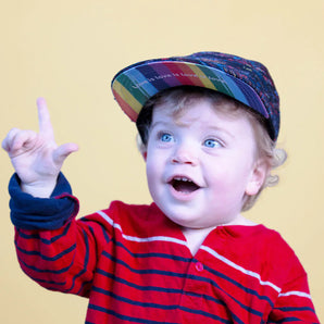 A happy toddler in a red striped shirt and Lil Hat by Storied Hats points up against a yellow backdrop.