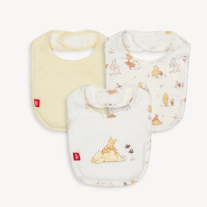 The Magnetic Me 3-Pack Bib Moments with Friends includes a yellow bib and two with Winnie the Pooh designs in playful poses, each featuring SewSafe™ magnetic closures and crafted from soft TENCEL™ modal. They all include a red tag with a white logo.