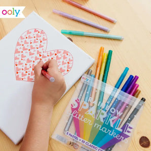 Someone uses OOLYs Rainbow Sparkle Glitter Markers to color a heart on paper, bringing vibrant charm to the wooden table.