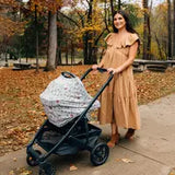 A woman in a long brown dress strolls through the park, pushing a stroller adorned with Milk Snobs Cover Disneys Winnie the Pooh Baby Bee cover. Autumn trees with vibrant orange and yellow leaves create a stunning backdrop.