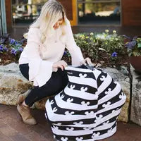 A woman sits on a stone bench outdoors, adjusting a Milk Snob Cover Disneys Mickey Mouse on a baby car seat. Her light pink blouse, black pants, and tan boots complement the vibrant flowers and building in the background.