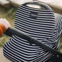 A close-up of a baby stroller features the Milk Snob Cover Modern Stripe nursing cover. A hand in an orange sleeve grasps the handle as the multifunctional cover fits snugly, framed by blurry greenery and a path.