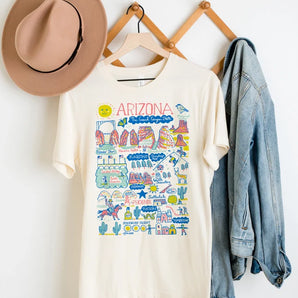 An Adult Organic Tee with an Arizona map by Morado Designs LLC hangs next to a denim jacket and a brown hat on a wooden rack.