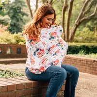 A woman sits outdoors on a brick ledge, using her Milk Snob Cover French Floral as a versatile nursing cover or baby car seat cover. Smiling and cradling her baby, she is surrounded by lush greenery and trees in the background.