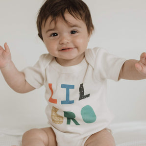 A smiling baby in Finn and Emmas Lil Bro organic bodysuit with raised arms features colorful, eco-friendly dyes.