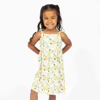 A young girl with long dark hair smiles brightly, wearing the Emerson and Friends Sundress in a vibrant Happy Honey Bees design. She stands confidently against a white background, hands on her hips.