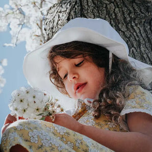 A young girl in a Jan and Jul white Sun Hat with chinstrap and floral dress sits in a tree, holding white flowers.