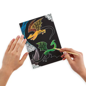 Hands use OOLYs Scratch & Scribble, Fantastic Dragon kit with a wooden stylus to reveal an orange and green dragon on black paper.