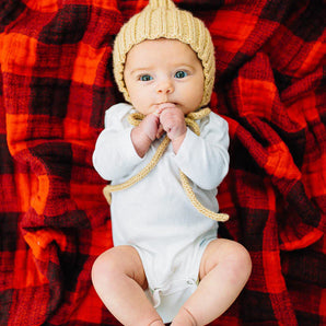 A baby with wide eyes lies under a Little Unicorn Cotton Swaddle, Red Plaid, wearing a knitted hat and white outfit.