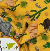 The Cabooties Arizona Animals feature a close-up design with yellow fabric adorned with cacti, cowboy hats, and brown shapes. An elastic ankle closure ensures a snug fit, while a round label with a green tree and star appears in the lower corner. Flexible rubber soles complete this baby shoe design.