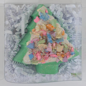 A green Christmas tree-shaped bath bomb with fun sparkles in clear packaging, nestled in white shredded paper.