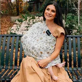 A woman sits on a park bench, smiling as she breastfeeds her baby under the Milk Snob Cover Disneys Winnie the Pooh Baby Bee. She wears a brown dress amid autumn leaves, and the multifunctional cover also serves as a chic accessory for her babys car seat.