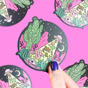 A hand with blue glitter nail polish holds the Desert Abduction vinyl sticker by Turtle Soup featuring cacti, coyotes, snakes, and skulls under a starry sky with a UFO shining down. The pink background displays the durable waterproof sticker repeatedly.