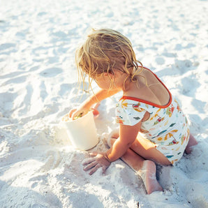 A curly-haired child in an Emerson and Friends One Piece Reversible Swimsuit plays with a white bucket on a sunny beach.