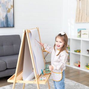 A girl with a bow in her hair paints at an easel, wearing the Bumkins Long-Sleeved Art Smock: Wander, in a cozy living room.