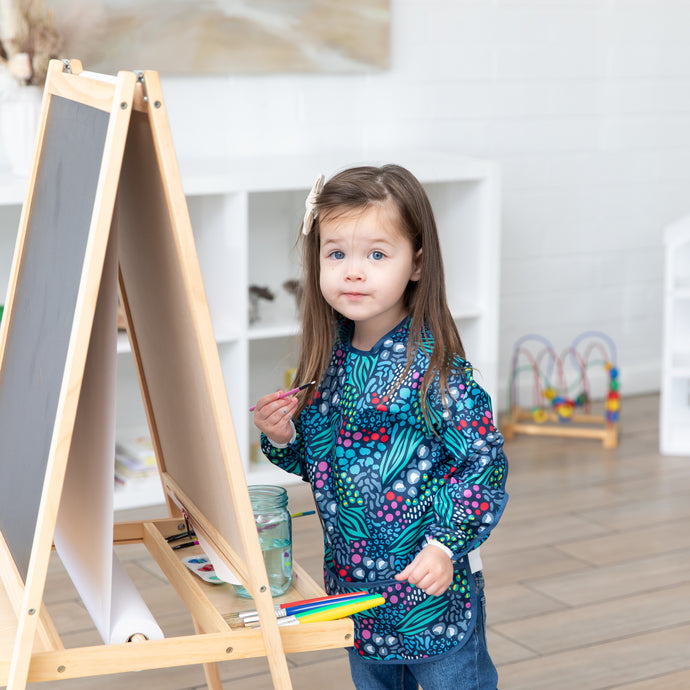 In a bright room, a young girl with long brown hair paints at an easel wearing the Bumkins Long-Sleeved Art Smock in animal prints. The adjustable closure protects her clothing, while art supplies and a bead maze toy scattered around inspire creativity without spill concerns.