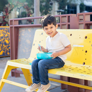 Young boy sitting on a yellow bench, smiling, and holding a Bumkins Silicone Bento Box Blue Jelly filled with food.