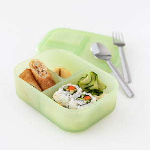 The Bumkins Silicone Bento Box, Green Jelly, crafted from LFGB-grade silicone, contains sushi rolls, vegetable salad, a spring roll, and a dumpling. A spoon and fork rest on the matching green lid against a plain white background.