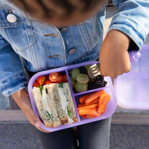 Child with Bumkins Silicone Bento Box in Purple Jelly, filled with a sandwich, fruits, and veggies. Wearing a denim jacket.