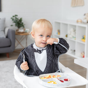 A toddler wearing a Bumkins Sleeved Bib in Jack Skellington enjoys food from a divided tray, sitting in a high chair with a cozy living room view.