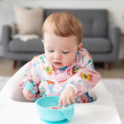 A baby sits in a high chair wearing a colorful bib, focused on a blue bowl in front. The baby is holding a spoon and appears to be feeding themselves. The background features a gray sofa.