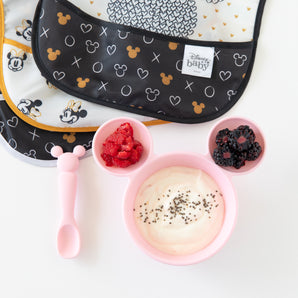 The Bumkins Minnie Mouse First Feeding Set includes a bib, Mickey bowl with yogurt, berries, and a pink spoon for baby self-feeding.