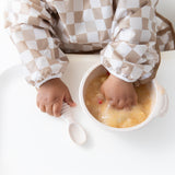 Young child in checker pattern explores mashed food with fingers from the Bumkins Silicone First Feeding Set: Sand, spoon nearby.