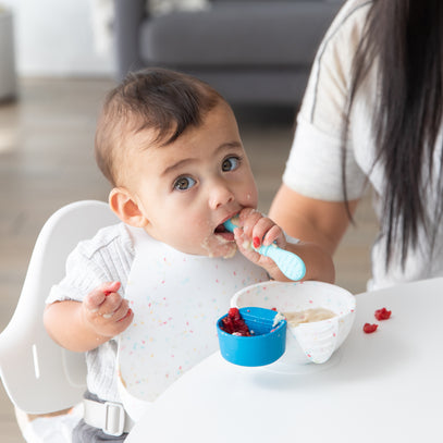 A baby sits in a high chair, wearing a bib, eating with a blue spoon. They have food around their mouth and hold a piece of food in one hand. A white bowl with bright-colored food is in front of them. An adult sits nearby.