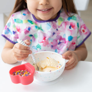 Child in a floral shirt beams while enjoying ice cream from a Bumkins Vanilla Sprinkle Silicone First Feeding Sets suction base bowl.