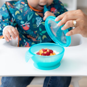 A child in a colorful bib enjoys fruit from the Bumkins Silicone First Feeding Set: Blue Jelly, as an adult holds the bowls suction lid.