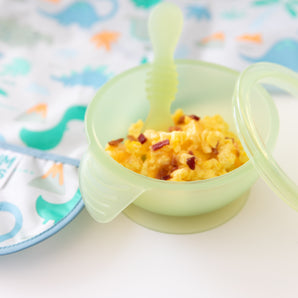 Baby bowl of scrambled eggs & bacon with a Bumkins Silicone First Feeding Set: Green Jelly and a colorful dinosaur-themed bib.