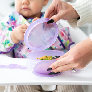 An adult opens a Bumkins Silicone First Feeding Set: Purple Jelly in front of a toddler in a floral bib, seated eagerly in their high chair.