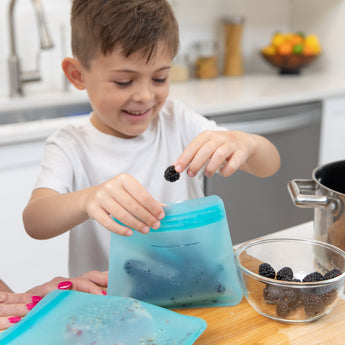 A smiling child in an eco-friendly kitchen places blackberries into a Bumkins Silicone Flat Reusable Bag, with a bowl nearby.