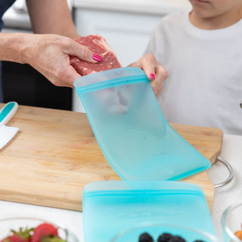 A person puts raw meat in a Bumkins Silicone Flat Reusable Bag as a child watches from the kitchen counter.