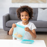 At the table, a child joyfully holds a Bumkins Silicone Flat Reusable Bag, Blue, with orange and lemon slices inside. With an orange slice in their mouth, they smile amid an eco-friendly scene featuring a gray sofa and white pillows in the background.