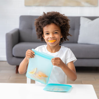 At the table, a child joyfully holds a Bumkins Silicone Flat Reusable Bag, Blue, with orange and lemon slices inside. With an orange slice in their mouth, they smile amid an eco-friendly scene featuring a gray sofa and white pillows in the background.