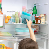 Person reaching into a fridge to grab a Bumkins Silicone Flat Reusable Bag, Blue. Eco-friendly kitchen items and drinks are inside.