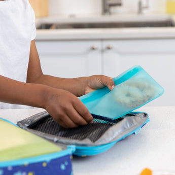 Packing a sandwich into a Bumkins Silicone Flat Reusable Bag, placing it in a blue lunchbox on the eco-friendly kitchen counter.