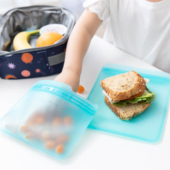 A childs hand reaches for snacks beside a sandwich as a Bumkins blue silicone bag, lunch bag, banana, and orange are in the background.