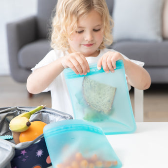 A young child seals a Bumkins Silicone Flat Reusable Bag, Blue, with a child-friendly zip seal around a sandwich. Seated at a white table with a lunchbox, banana, and orange nearby, the scene suggests an eco-friendly kitchen setting for packing lunch in the living room.